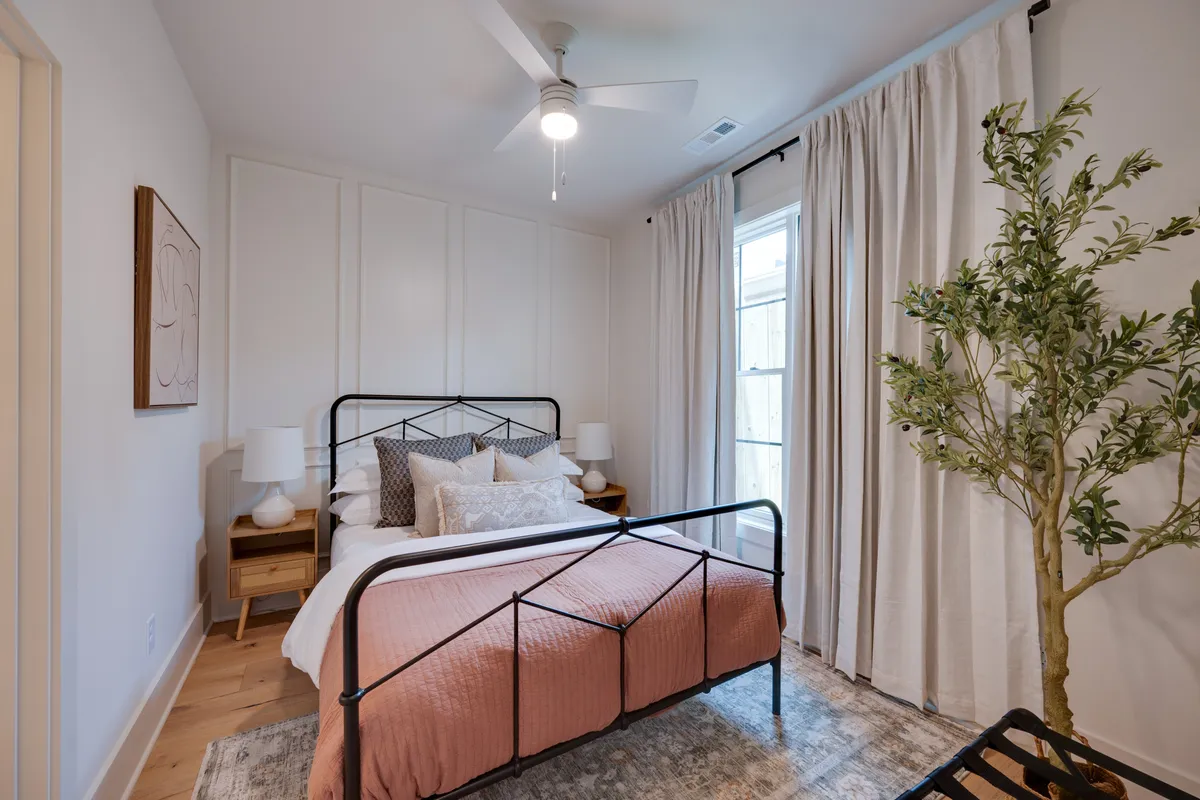 A cozy queen-bedded room with soft pink bedding, decorative pillows, and abundant natural light streaming through the window. Nightstands with lamps and an indoor plant add to the intimate, calming ambiance—suggesting a restful space with thoughtful amenities.