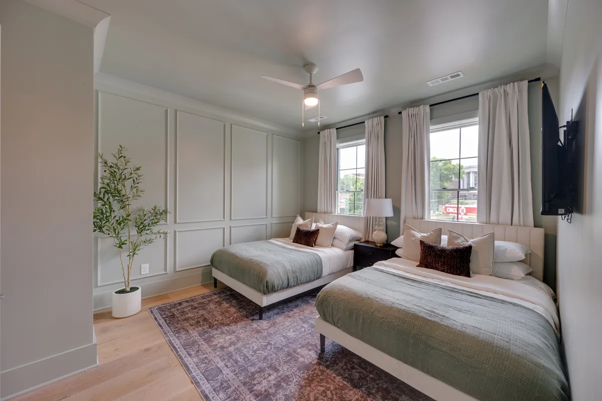 A bright and tranquil bedroom with two full beds topped with plush bedding and accent pillows. Large windows flood the room with natural light, while decorative plants and soft furnishings enhance the sense of comfort. The arrangement is inviting—perfect for families or friends sharing a room.