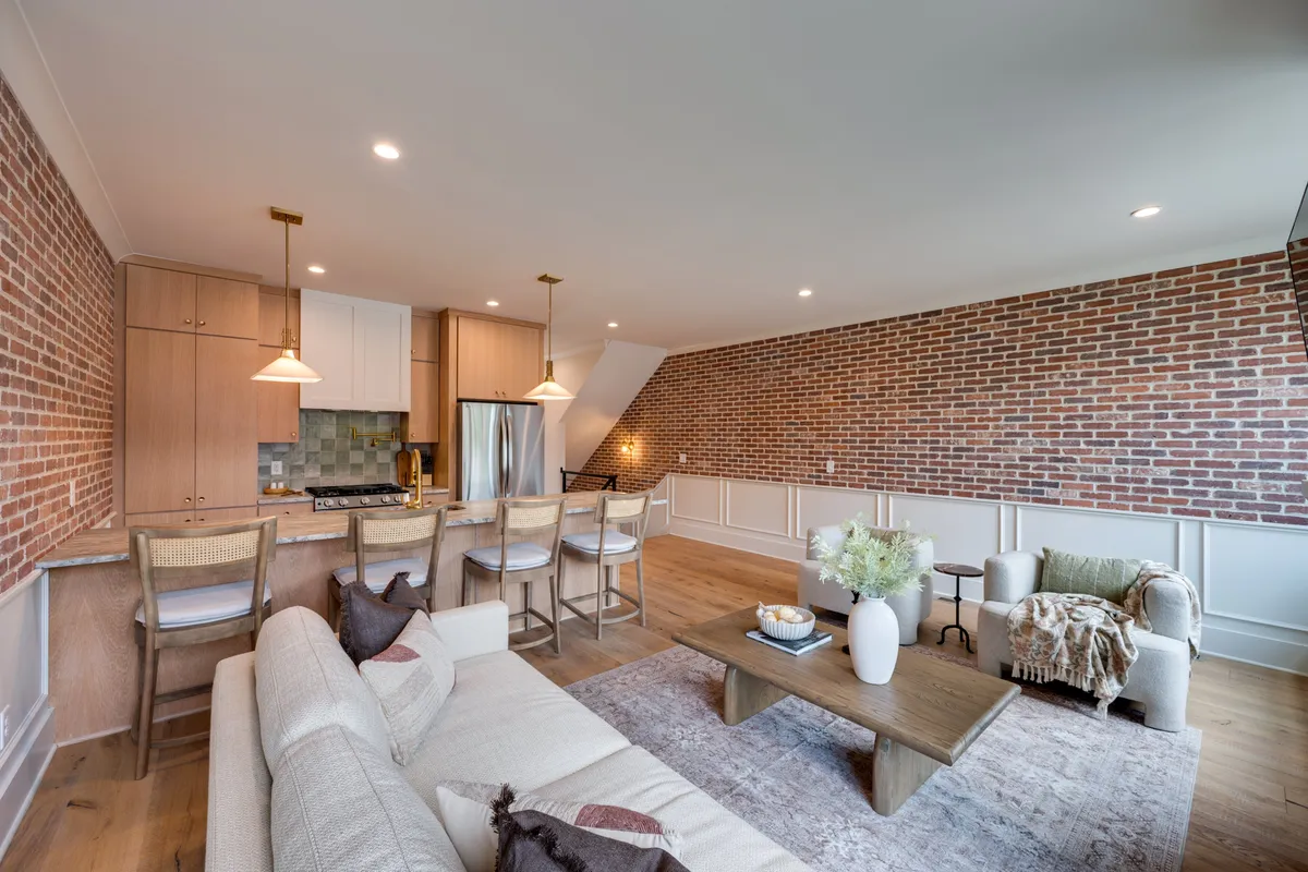 The image showcases a beautifully designed interior space featuring an exposed brick wall that adds historic Nashville charm, a sleek kitchen island perfect for gatherings, a plush and inviting sofa, and a warm wooden coffee table. The layout creates a seamless flow for socializing, dining, and relaxing, all bathed in natural light.