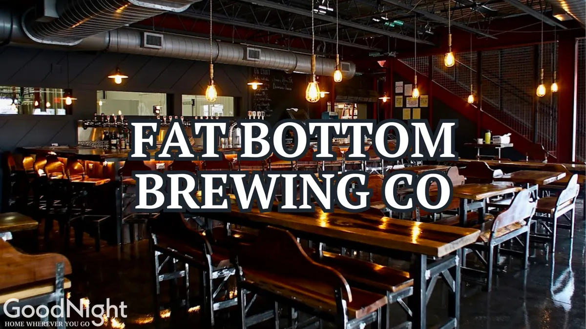 An open-plan interior featuring long wooden tables, a well-lit bar area, hanging light fixtures, and an exposed brick wall. The setting is designed for craft beer tasting, social gatherings, and casual dining, radiating a relaxed and communal vibe.
